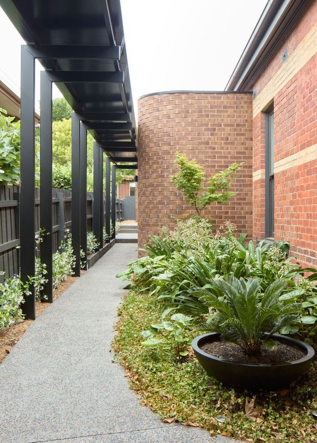 A narrow landscaped external walkway runs beside a red brick building. A black steel pergola or covered structure with regularly spaced posts lines the left side, while a curved brick wall and planted garden bed occupy the right. The path has a light grey textured paving finish, with dense greenery and a large black planter in the foreground. The image has a contemporary architectural landscape character with strong linear framing and contrasting dark steel and warm brick.