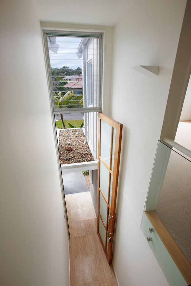 A narrow, bright stairway or landing viewed from above, with white walls, a tall glazed window, and a timber-framed glass door opening toward an exterior entry path. A glass balustrade with a timber handrail borders the stair opening on the right. Light wood-toned flooring and neutral finishes create a clean, airy interior, with suburban greenery visible outside.