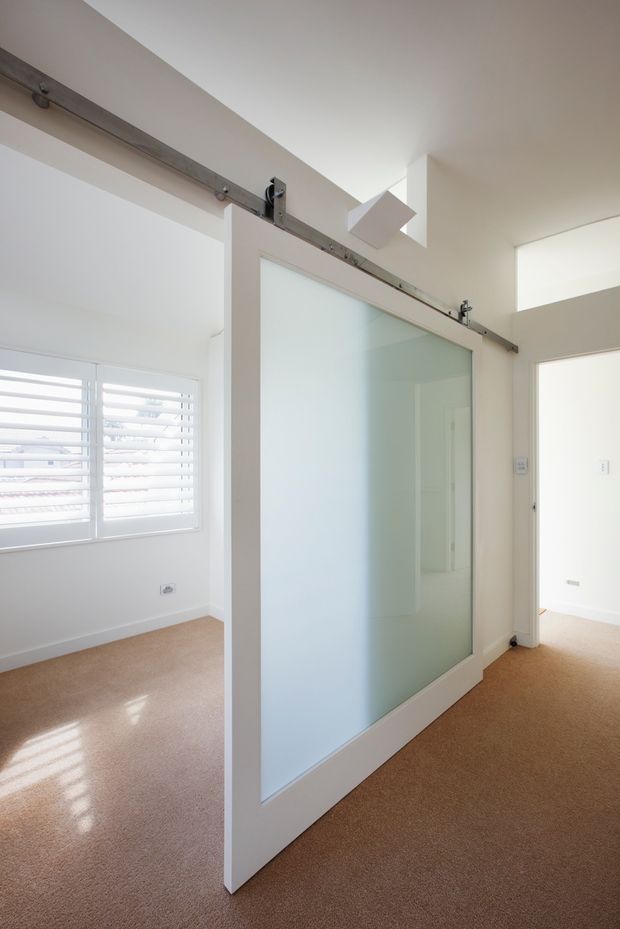 Bright minimalist interior hallway or room divider featuring a large white-framed frosted-glass barn-style sliding door on exposed metal hardware. White walls and ceiling, a high ceiling volume with clerestory-like glazing, plantation shutters, and warm beige carpet create a clean, airy space.