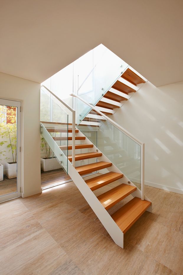 Bright contemporary interior featuring a sculptural open-riser staircase with warm timber treads, white stringers, and clear glass balustrades. The staircase rises through a high angular ceiling void, with pale timber flooring, white walls, and abundant natural light.
