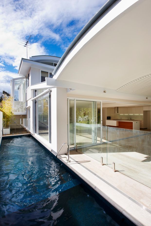 Bright exterior view of a contemporary white multi-level home with broad roof overhangs, extensive glazing, a narrow swimming pool alongside the building, and an open-plan kitchen visible through sliding glass doors. The pool deck and indoor floor continue visually across the fully glazed opening.