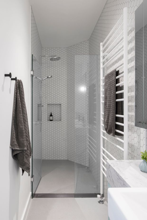 Bright, narrow contemporary bathroom with a walk-in glass shower enclosure. The shower walls are clad in small white hexagonal tiles, with a recessed niche, dual shower fittings, and a grey tiled floor. A white heated towel rail with dark towels occupies the right wall beside a marble-look vanity benchtop and mirrored cabinet. The palette is predominantly white and light grey with restrained dark accents.