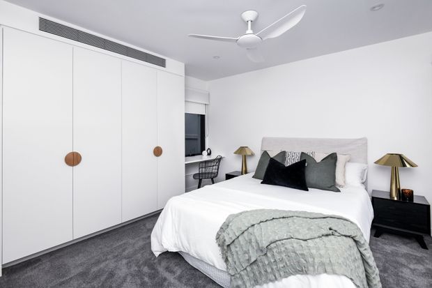 A bright, contemporary bedroom with a large white built-in wardrobe featuring round timber pulls, a neatly made bed with layered grey and black cushions, matching bedside tables and lamps, grey carpet, and a white ceiling fan. A compact desk is positioned near the window.