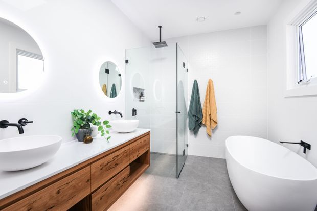 Bright contemporary bathroom with a double vanity featuring warm timber cabinetry and white vessel basins, a walk-in glass shower, freestanding white bathtub, large-format pale wall tiles, grey floor tiles, black tapware and fittings, and round mirrors. Natural light enters through the high side window, while towels and a potted plant add subtle colour and texture.