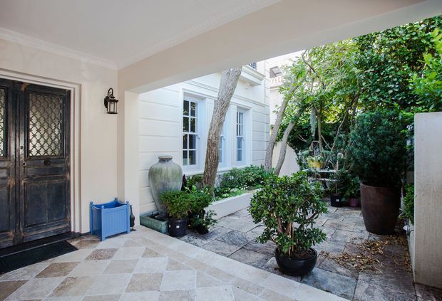 A refined covered entry courtyard with a dark decorative timber front door, pale rendered walls, tiled flooring, and a landscaped garden with potted shrubs and trees. White-framed French doors and a light-coloured rendered building are visible beyond the courtyard, creating a quiet, leafy arrival space.