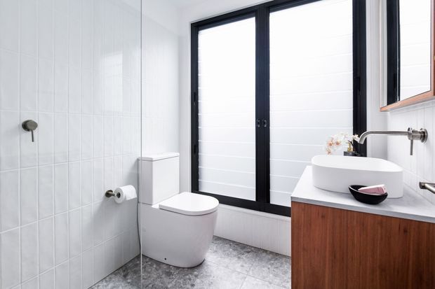 Bright contemporary bathroom with white vertical wall tiles, a wall-mounted shower fixture, a sculptural white toilet, black-framed full-height glazing with horizontal privacy screening, and a floating timber vanity with a pale benchtop and vessel basin. The space has a clean, high-contrast palette with warm timber joinery.