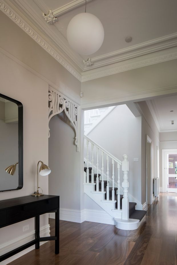 A refined traditional hallway and stairway with ornate plaster ceiling cornices, decorative wall arch detailing, white turned stair balusters, dark carpeted stairs, and polished dark timber flooring. A black console table, framed mirror, and small table lamps sit along the left wall, while a bright corridor extends toward the rear of the home.