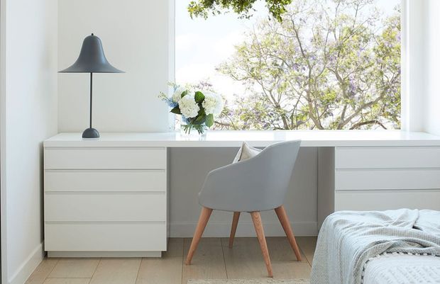 A bright, minimalist bedroom workspace set beneath a large window. A white built-in desk with drawer units spans the window wall, paired with a pale upholstered chair on timber legs. A slim black table lamp, vase of flowers, and neatly made bed with a light textured throw complete the calm, neutral interior.