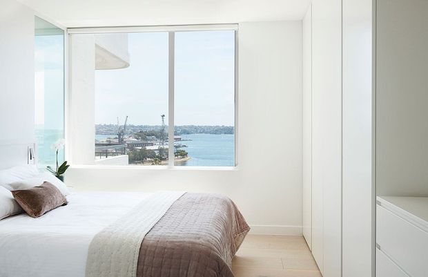 Bright, minimalist bedroom with a neatly made bed, pale timber flooring, white walls and built-in white cabinetry. A large window provides expansive water views, while soft neutral bedding and a small potted orchid add understated warmth.