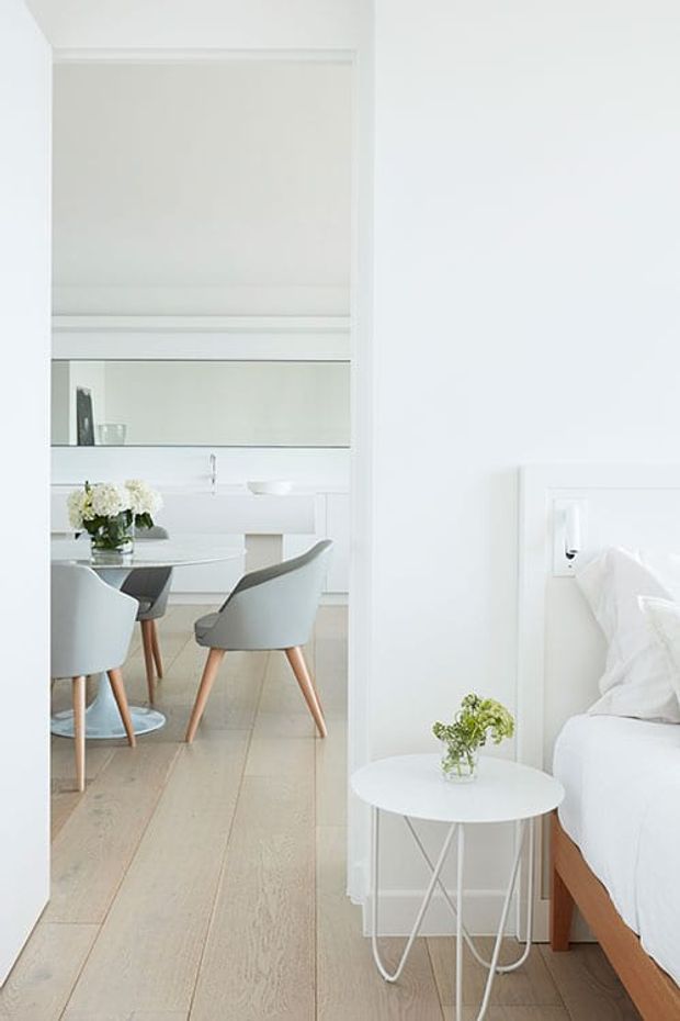 Bright, predominantly white bedroom viewed through an open doorway, with a partially visible bed, white bedside table, and small floral arrangement. Beyond the bedroom is a light-filled dining area with a round table, upholstered dining chairs, pale timber flooring, and built-in white cabinetry. The space has a clean, restrained, minimalist appearance with soft natural light.