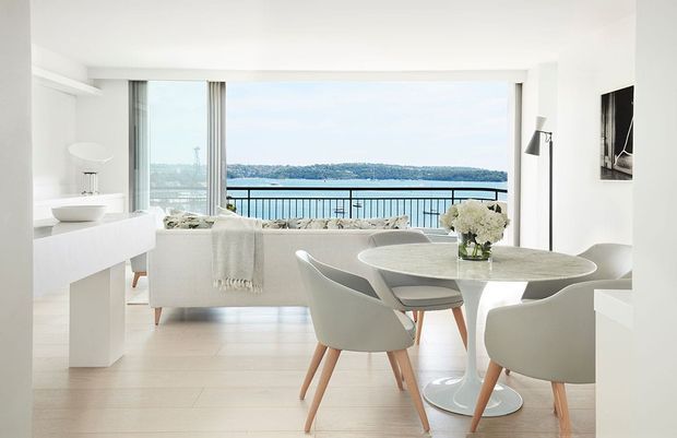 Bright, open-plan apartment interior combining a dining area and living room, with white contemporary furnishings, pale timber flooring, a sculptural round dining table, and full-height glazing opening to a balcony with water views. The space has a clean, airy coastal feel and abundant natural light.