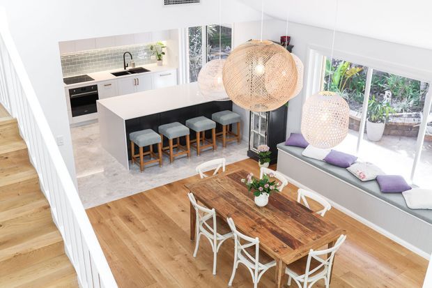 Bright, open-plan kitchen, dining, and living area viewed from an elevated stair or landing. The room features a white kitchen with a large island and seating, pale stone-look flooring, warm timber flooring in the dining zone, a rustic timber dining table with white chairs, a window seat, and large woven pendant lights. Abundant glazing and indoor plants create an airy, contemporary coastal feel.