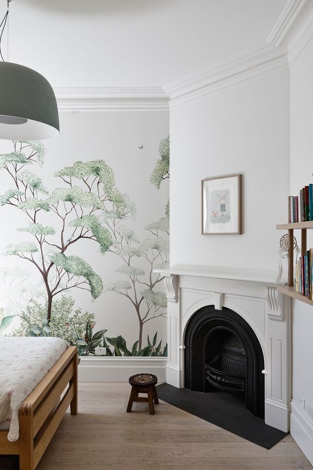 A bright, refined bedroom corner with a timber bed partially visible at left, pale engineered timber flooring, and a traditional white-painted fireplace with a black cast-iron insert. The walls feature a delicate botanical tree mural, while built-in open shelving displays books and small decor. Ornate ceiling cornices, wall mouldings, and fireplace detailing add a classic architectural character, complemented by a large grey pendant light and framed artwork.