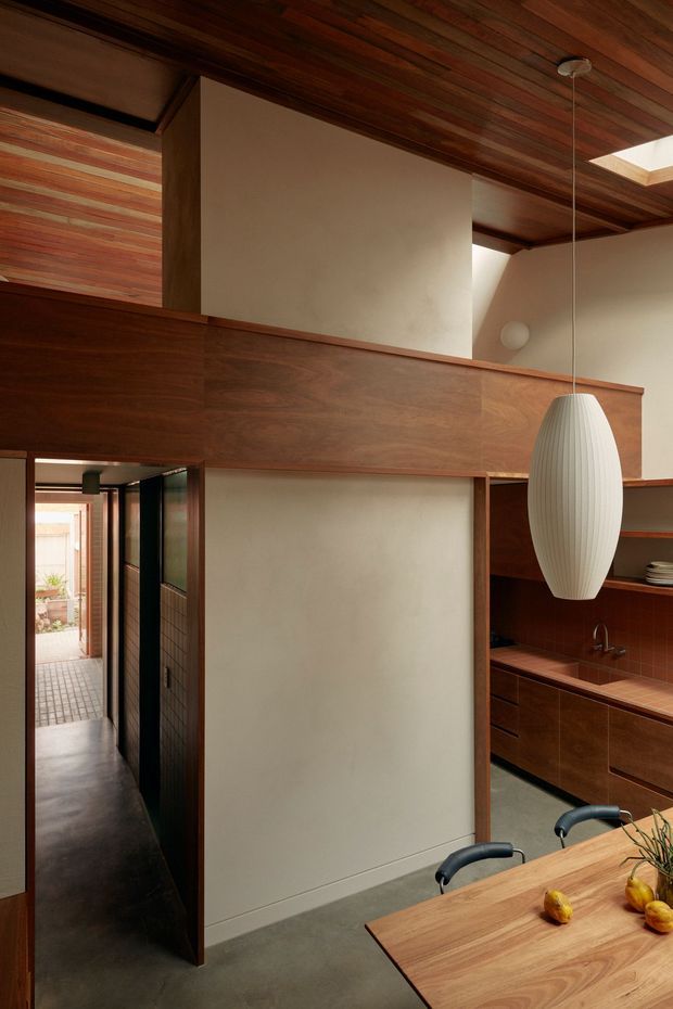 Interior view of a warm, wood-lined open-plan kitchen and dining area. Dark timber wall panelling and ceiling boards frame pale plaster walls, with a long horizontal timber partition creating a mezzanine-like upper zone. A sculptural white pendant hangs above a timber dining table, while the compact kitchen features dark wood cabinetry, a reddish tiled benchtop and splashback, open shelving, and a sink. A narrow hallway leads toward a small courtyard or exterior walkway.