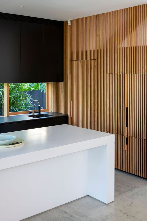 Contemporary kitchen interior featuring a large white island with a waterfall-style benchtop, matte black cabinetry and sink fixtures, polished concrete flooring, and a full-height vertical timber slat feature wall concealing integrated storage. A window above the rear bench overlooks lush planting.