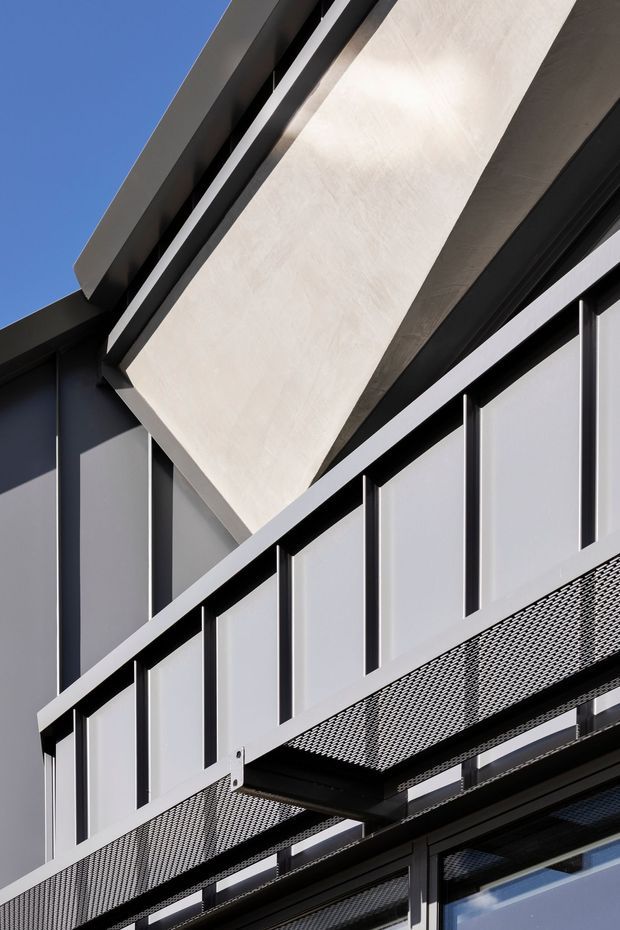 Close-up upward view of a contemporary building facade featuring dark metal cladding and framing, pale angular projecting panels or awnings, a perforated metal balcony or walkway edge, and large glazed openings. The image emphasizes sharp geometric forms, layered projections, and contrasting light and dark architectural materials against a clear blue sky.