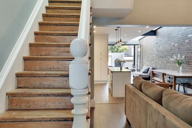 Interior view from a stairway landing looking toward an open-plan kitchen, dining, and living area. The foreground features a traditional timber staircase with a turned white balustrade, while the bright renovated space beyond combines light cabinetry, timber flooring, a central island, dining furniture, and an exposed grey brick feature wall.