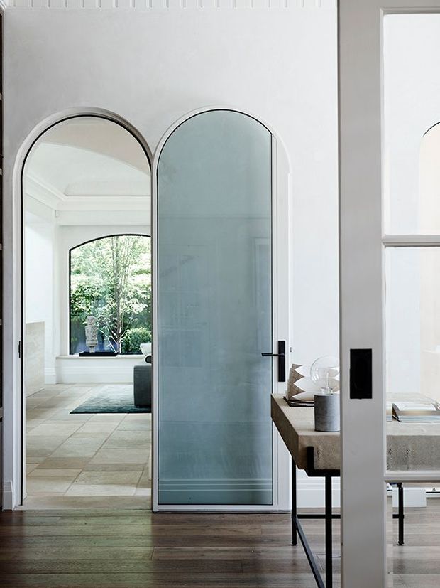 A refined interior view centered on a tall arched glazed door with a slim dark frame and black hardware. The door separates a dark timber-floored room from a bright tiled sitting or living area with an arched window, greenery visible beyond, and a low rug. To the right is a minimalist console or desk with a stone-like top and dark metal legs, alongside white-framed glazing and arched architectural detailing. The palette is predominantly white, pale grey, light timber, and black accents.