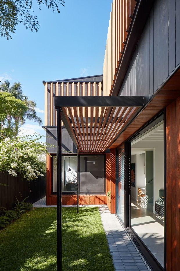 A bright exterior view of a contemporary residential courtyard or garden-facing passage. The house features dark vertical cladding, warm timber wall lining and vertical timber screening, large sliding glass doors, and a projecting pergola with closely spaced timber slats supported by slender dark metal posts. A paved walkway runs alongside a small lawn and planted garden, with interior living space visible through the open glazing.