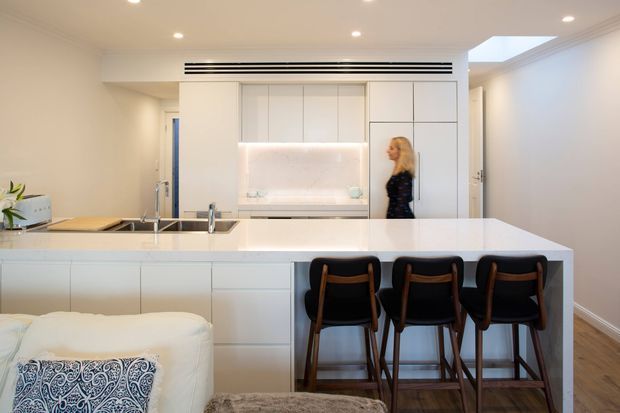 Bright, refined open-plan kitchen viewed from an adjoining living area. A large white island with integrated sink and three dark timber-and-black bar stools dominates the foreground. The kitchen features extensive flat-panel white cabinetry, a pale illuminated stone-look splashback, built-in appliances, recessed ceiling downlights, linear black air vents, and a skylight. Timber flooring continues through the space, while a white sofa and patterned cushion are visible in the foreground.