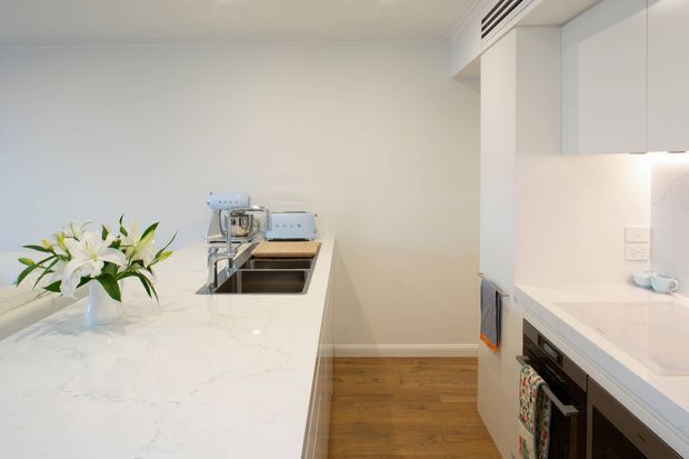Bright, compact contemporary kitchen with white cabinetry and white marble-look benchtops. A long peninsula or island with an inset double sink and chrome tap dominates the foreground, while integrated appliances and a cooktop are visible along the right wall. Warm timber flooring contrasts with the predominantly white fit-out, and a vase of lilies provides a small decorative accent.