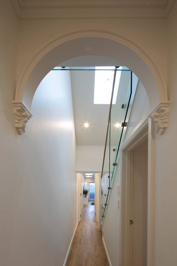 A narrow, elongated interior hallway viewed through an ornate arched opening. The corridor has warm timber flooring, white walls and ceiling, recessed downlights, a roof skylight, and a modern clear glass balustrade with slim metal fittings along the right side. Decorative classical mouldings and sculpted corbels frame the arch and doorway, while several rooms open off the hall toward a distant glazed door.