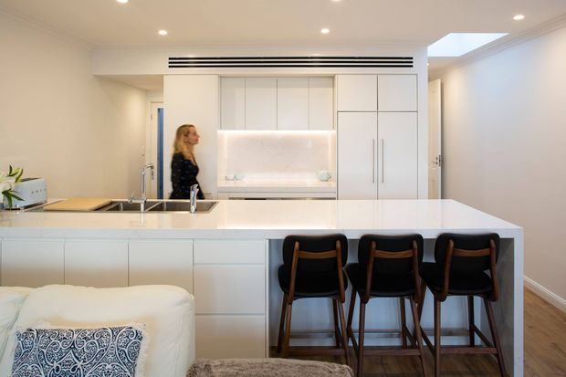 Bright, contemporary open-plan kitchen with a large white island and three dark timber bar stools. The space features extensive white flat-panel cabinetry, integrated appliances, a softly illuminated backsplash or display niche, recessed ceiling lights, timber flooring, and a connected living area in the foreground.