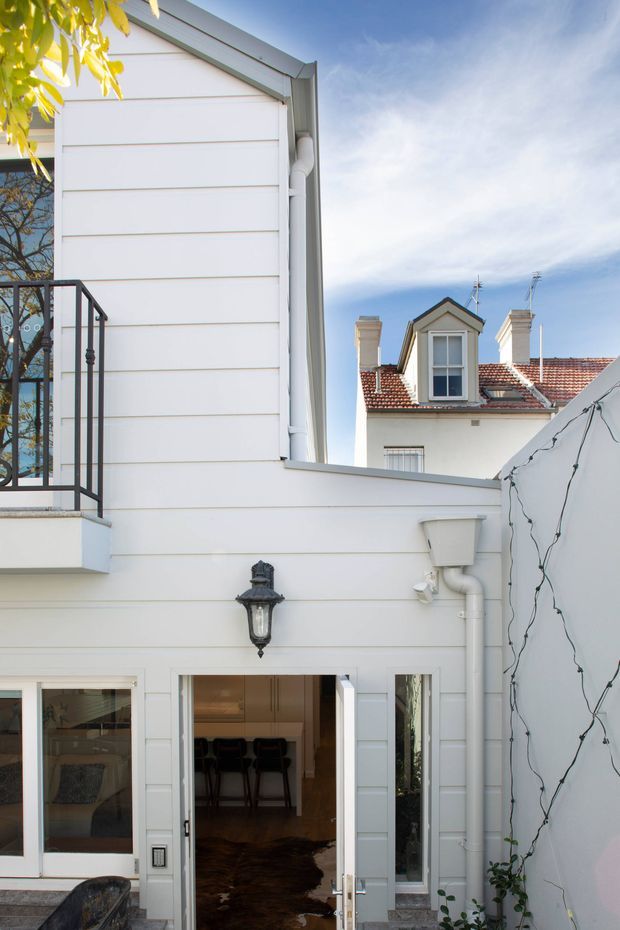 A bright, narrow urban courtyard viewed from outdoors, enclosed by a white multi-level residential facade. The foreground building has horizontal white weatherboard-style cladding, a black metal balcony railing, white-framed glazed doors and windows, a traditional wall lantern, and exposed white drainage pipes. A neighboring heritage-style building with a tiled roof, chimneys, and dormer window is visible beyond the courtyard. String lights run along the right-hand wall, while an interior kitchen or bar area is visible through the open doorway.