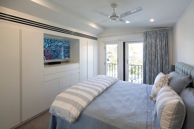 Bright, compact bedroom with a built-in wall of white cabinetry and integrated media niche, a low-profile bed dressed in layered pale blue and white bedding, and upholstered grey-blue headboard. French doors open to a small balcony, while patterned curtains, recessed ceiling lights, and a ceiling fan complete the clean coastal-inspired interior.