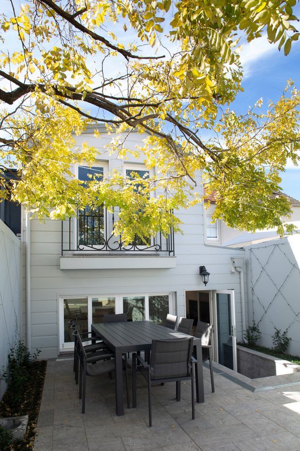A sunlit outdoor courtyard or patio viewed toward the rear facade of a two-storey home. The white horizontal-clad exterior has upper-level windows with decorative black wrought-iron balustrades and lower-level glazed doors opening onto the paved entertaining area. A dark outdoor dining table and chairs occupy the foreground, with mature leafy branches overhead and a raised planter along the side wall.