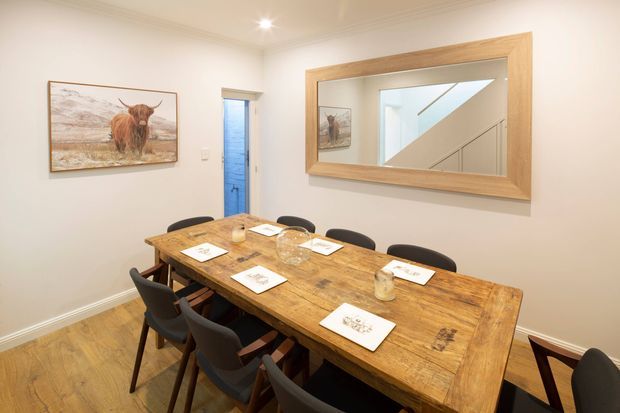 A bright, compact dining room with a large rustic timber dining table surrounded by dark upholstered chairs. White walls, timber flooring, a broad timber-framed wall mirror, recessed ceiling lights, framed artwork, and a glimpse of a stairwell through an open doorway create a warm, contemporary-traditional interior.
