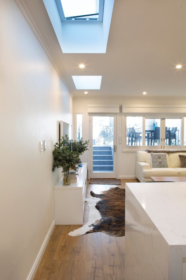 Bright open-plan living and kitchen area viewed toward a glazed rear door and windows. The room has white walls with decorative cornice detailing, recessed downlights, two large skylights, warm timber flooring, a white built-in kitchen island with a pale stone benchtop, a cream sofa, and a compact white media console. A cowhide rug and potted greenery add contrast. Outdoor dining furniture is visible through the glazing.