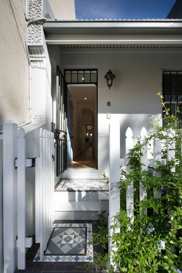 A narrow residential entryway viewed from outside, approached by tiled steps and framed by a white picket gate and fence. The facade features pale painted cladding, black-framed glazing, a covered porch, decorative wall lantern, and house number 6. Patterned geometric floor tiles add a strong decorative element, while leafy planting partially screens the entrance. The image has a bright, refined period-inspired urban character.