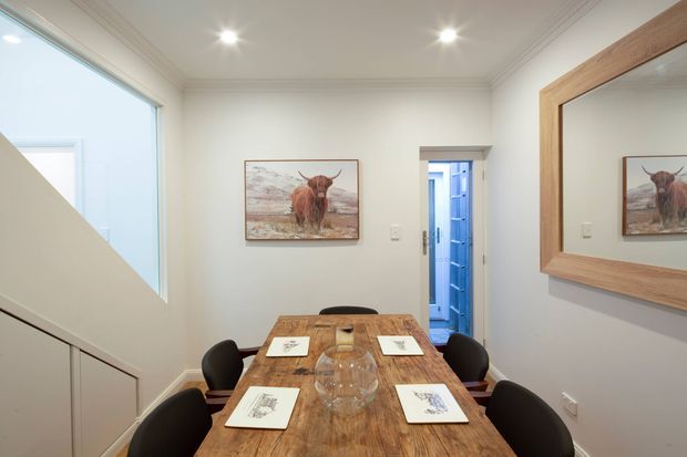 Compact dining room with a rustic timber dining table surrounded by dark upholstered chairs. The room has white walls and ceiling with decorative cornice, recessed ceiling lights, a large timber-framed wall mirror, and a highland cattle artwork. A glazed stairwell or landing is visible at left, while a doorway opens to a blue-toned adjacent area.