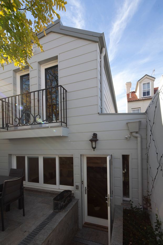 Exterior view of a compact, multi-level white weatherboard-clad building enclosing a paved courtyard or terrace. The facade has tall glazed doors, a small upper balcony with decorative black metal railing, white-framed windows and doors, and a traditional wall lantern. A neighbouring dwelling with a dormer window and red tiled roof is visible beyond the courtyard.