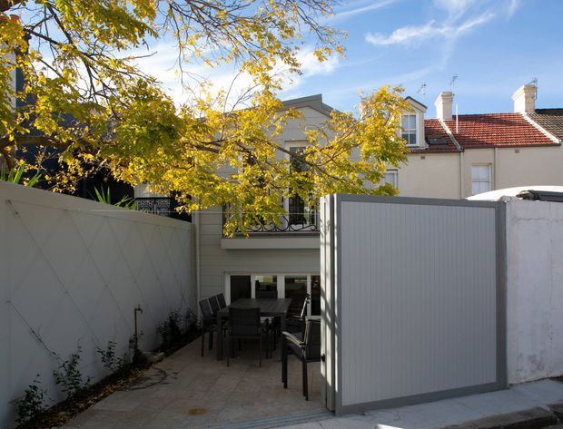 A sunny enclosed residential courtyard or rear outdoor dining area, viewed from the street through an open grey metal gate. The paved terrace contains an outdoor dining table and chairs, with tall light-coloured boundary walls, a black metal balcony railing, and a cream rendered multi-storey house with a red tiled roof and chimneys behind it. Mature leafy branches partially frame the scene.
