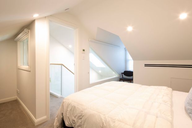 A bright, compact attic bedroom with a sloping ceiling, white walls and trim, recessed ceiling lights, and a large white bed. An open doorway leads to a stair landing with a glass balustrade, while an angled skylight or dormer window provides natural light. A small dark chair sits beside the window, and beige carpet covers the floor.