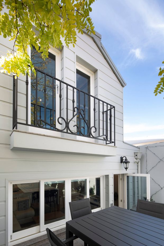 Exterior view of a two-storey white weatherboard-clad dwelling with a steep gable roof, tall windows, and a projecting upper-level balcony. The balcony has ornate black metal balustrades, while the lower level opens onto a furnished outdoor dining terrace with a dark table and chairs. A traditional wall lantern and climbing vegetation add character.