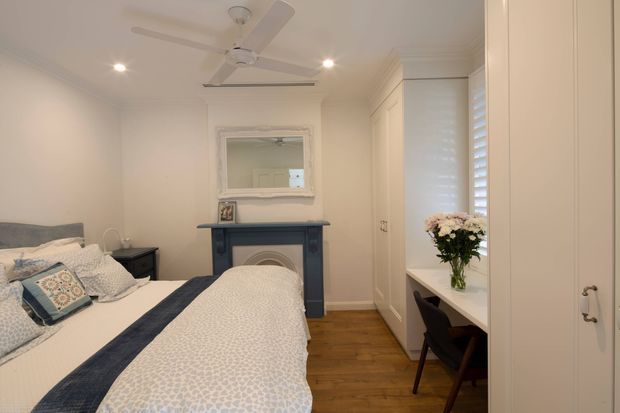 A bright, compact bedroom with two single beds dressed in pale patterned bedding and navy runners. The room features white built-in wardrobes, a small integrated desk beneath plantation shutters, a dark decorative fireplace mantel with a mirror above, warm timber flooring, recessed ceiling lights, and a white ceiling fan. The overall styling is classic and restrained with blue-and-white accents.