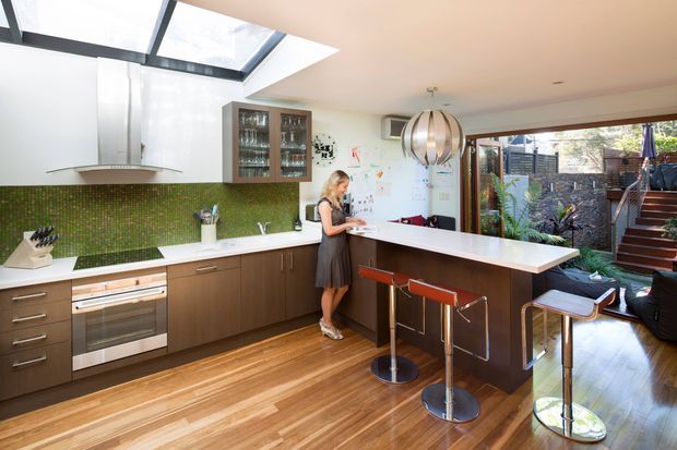 A bright contemporary open-plan kitchen with a long white island and breakfast bar, dark wood cabinetry, a green mosaic tile splashback, stainless-steel appliances, and warm timber flooring. A large skylight brings in natural light, while full-height glazing opens toward a planted outdoor courtyard and exterior stairway. Glass-fronted display cabinetry and a sculptural pendant light add visual interest.