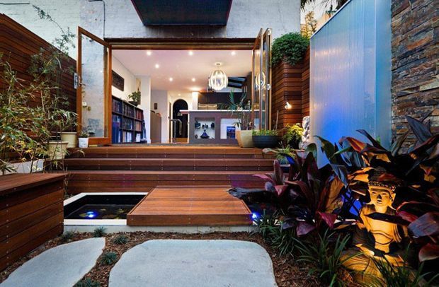 A landscaped entrance courtyard leading into a modern home through wide timber-framed glazed doors. The approach features broad timber steps and decking, large pale concrete stepping stones, a small rectangular water feature, lush planting, illuminated sculptural garden decor, timber privacy screens, a rendered wall, and a stacked-stone feature wall. The interior beyond shows shelving, a compact bar or display joinery, and contemporary lighting.