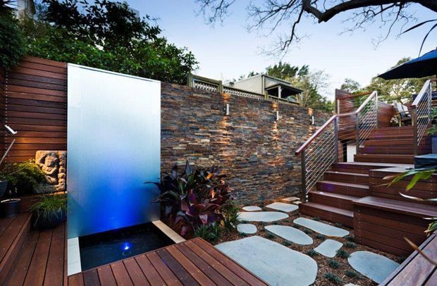 A landscaped outdoor courtyard with timber decking, a feature water wall and basin, layered natural-stone retaining wall, and broad stepping stones leading to raised timber stairs. Metal cable balustrades and dense planting create a private, contemporary garden setting.