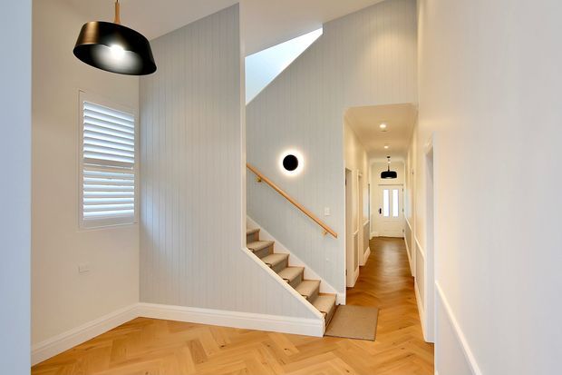 Bright residential hallway and stairway with warm herringbone timber flooring, white vertical wall panelling, white trim, a compact staircase with timber handrail, shuttered window, and contemporary pendant and wall lighting. The corridor extends toward a glazed front door, creating a clean, refined transitional space.