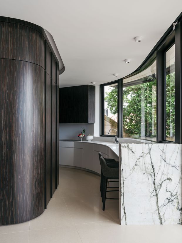 A refined contemporary kitchen with a gently curved plan and dark ribbed timber cabinetry forming a dramatic rounded wall. The space features a pale stone floor, curved grey cabinetry, black-framed windows, integrated lighting, and a large veined white marble or natural-stone island. A small seating area sits at the island, with abundant greenery visible outside.