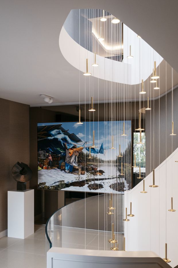 A dramatic contemporary interior stairwell with a sculptural curved void in the ceiling, a sweeping white staircase, glass balustrade, and a large installation of many slender pendant lights suspended at different heights. Dark textured walls contrast with white architectural surfaces, while a large artwork or digital display occupies the landing area. A sculptural object sits on a white plinth.