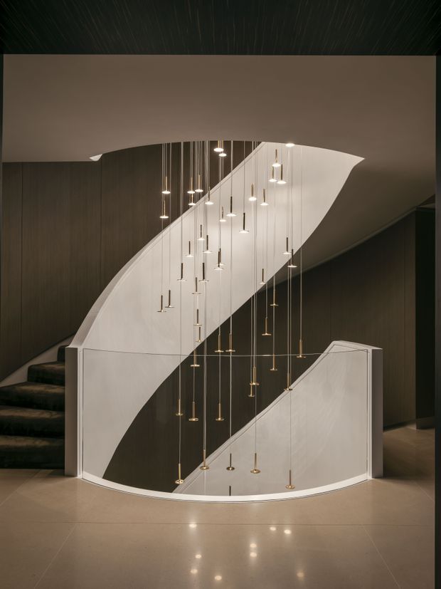 A dramatic contemporary stairway with sculptural curved white balustrades enclosing a dark stairwell. Numerous slender pendant lights hang at varying heights through the void, creating a refined focal feature. Dark timber or wood-look wall panelling contrasts with the pale polished floor and white stair surfaces.