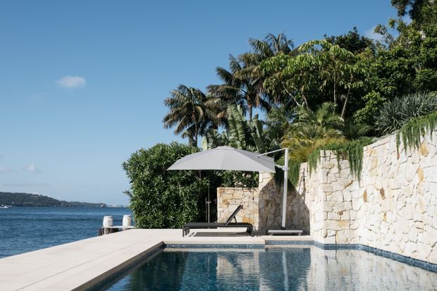 A sunlit waterfront pool terrace with a narrow rectangular swimming pool beside a pale natural-stone retaining wall. A light timber deck projects toward calm blue water, with loungers, a large umbrella, dense tropical planting, and palm trees surrounding the space.
