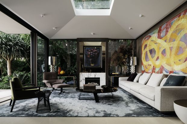 A sophisticated contemporary living room with floor-to-ceiling glazing on three sides, a central skylight, fireplace feature wall, large sectional sofa, sculptural coffee table, lounge chairs, and a bold abstract artwork. The room opens visually to dense greenery outside and is finished in a refined neutral palette with dark accents.