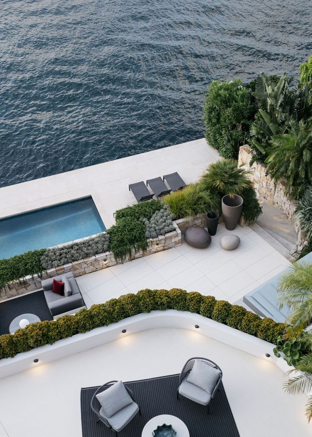 Elevated view of a luxurious multi-level waterfront outdoor area with a rectangular swimming pool, pale tiled terraces, dark outdoor lounge furniture, sculpted hedges, tropical planting, stone retaining walls, and a lower curved terrace overlooking the water.