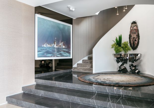 A sophisticated residential entry foyer with broad tiered dark stone steps, a curved staircase with carpeted treads, and a sculptural white curved wall. The space features dark reflective stone flooring and risers, a large framed artwork, mirrored wall panels, a geometric console table, a round organic-patterned rug, decorative greenery, and a carved wall mask. Recessed and pendant lighting create a refined, contemporary atmosphere.